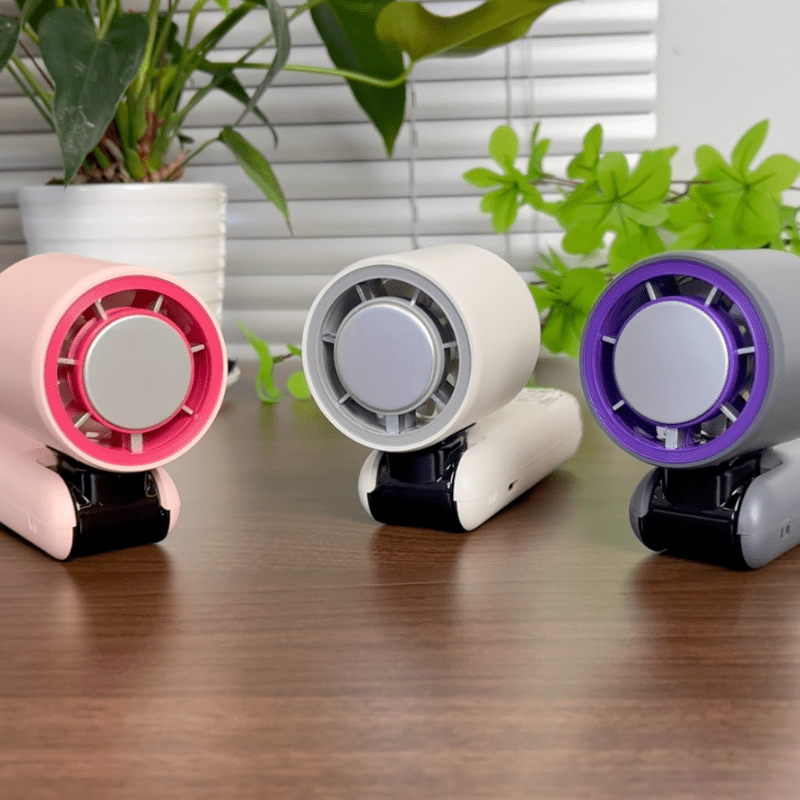 Three handheld turbo fans in pink, white, and grey with cooling system displayed on a wooden desk with indoor plants - techynook